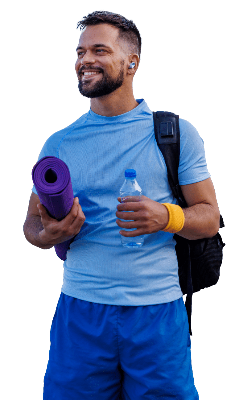 Man stands by the riverside, ready for a workout with a rolled-up yoga mat, water bottle, and a backpack. Dressed in a blue shirt and yellow wristband, he looks invigorated.